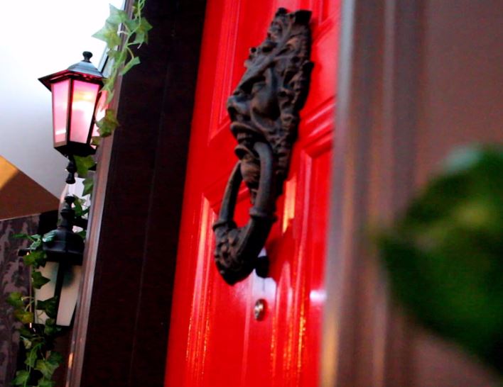 Red door with ornate black knocker next to vine-covered lantern.