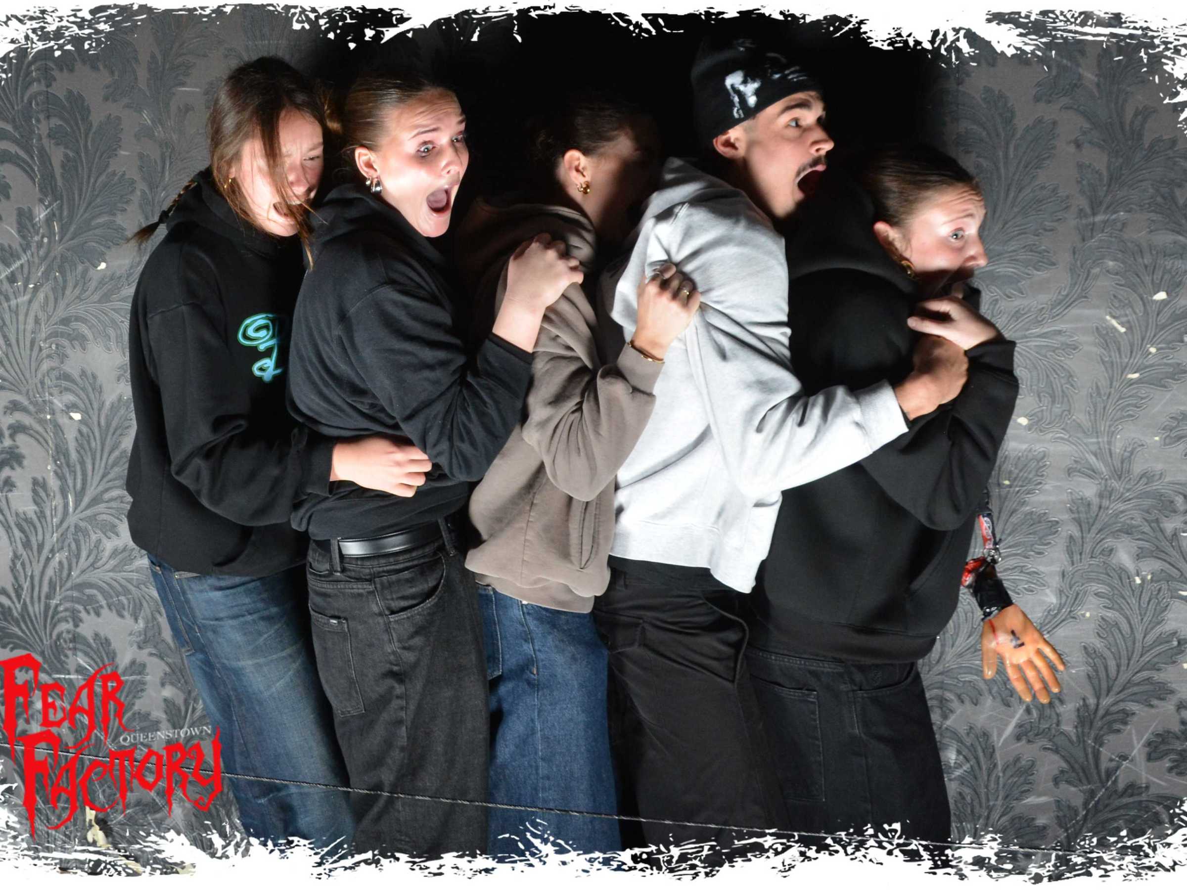 Five people in a dark room look scared, huddled together, wearing casual clothes.